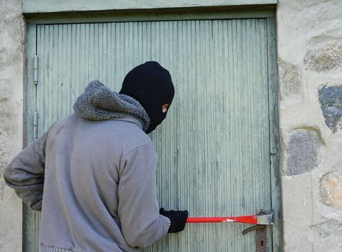 Mann mit grauem Pulli und Sturmmaske versucht eine Tür aufzuhebeln mit roter Brechstange