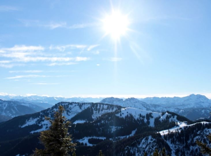 Blick vom Brecherspitz auf leicht mit Schnee bedeckten Berggipfeln, Fichtenwipfel im Vordergrund, blauem Himmel und gleißen heller Sonne