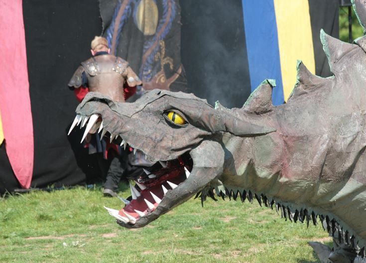 Große Drachenfigur mit aufgerissenem Maul schaut nach links. Im Hintergrund mittelalterliches Zelt, indem gerade ein Ritter eintritt