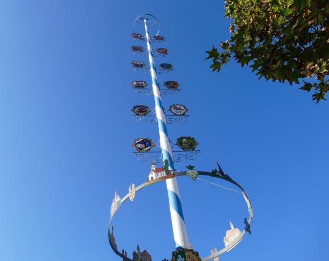 Spitze eines Maibaums mit blauem Himmel im Hintergrund. Links und rechts am Maibaumstangerl viele Figuren und unten am Bildrand ein Kranz aus Metall drum herum.