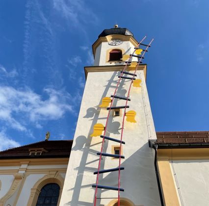 Blick zum Kirchturm hinauf in den blaue-weissen Himmel. Am Kirchturm hinauf führt das Kunstprojekt "Himmelsleiter" mit kleinen, gelben Flügelchen