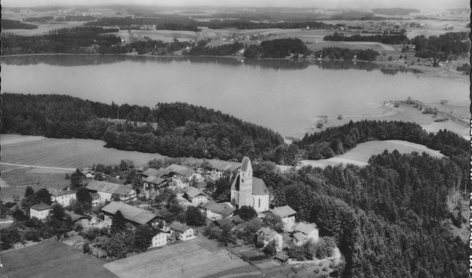 Schwarz-Weiss-Luftaufnahme des Orts Hirnsberg. Im Mittelpunkt der Kirchturm. Hinter dem Ort Bäume und dann der See