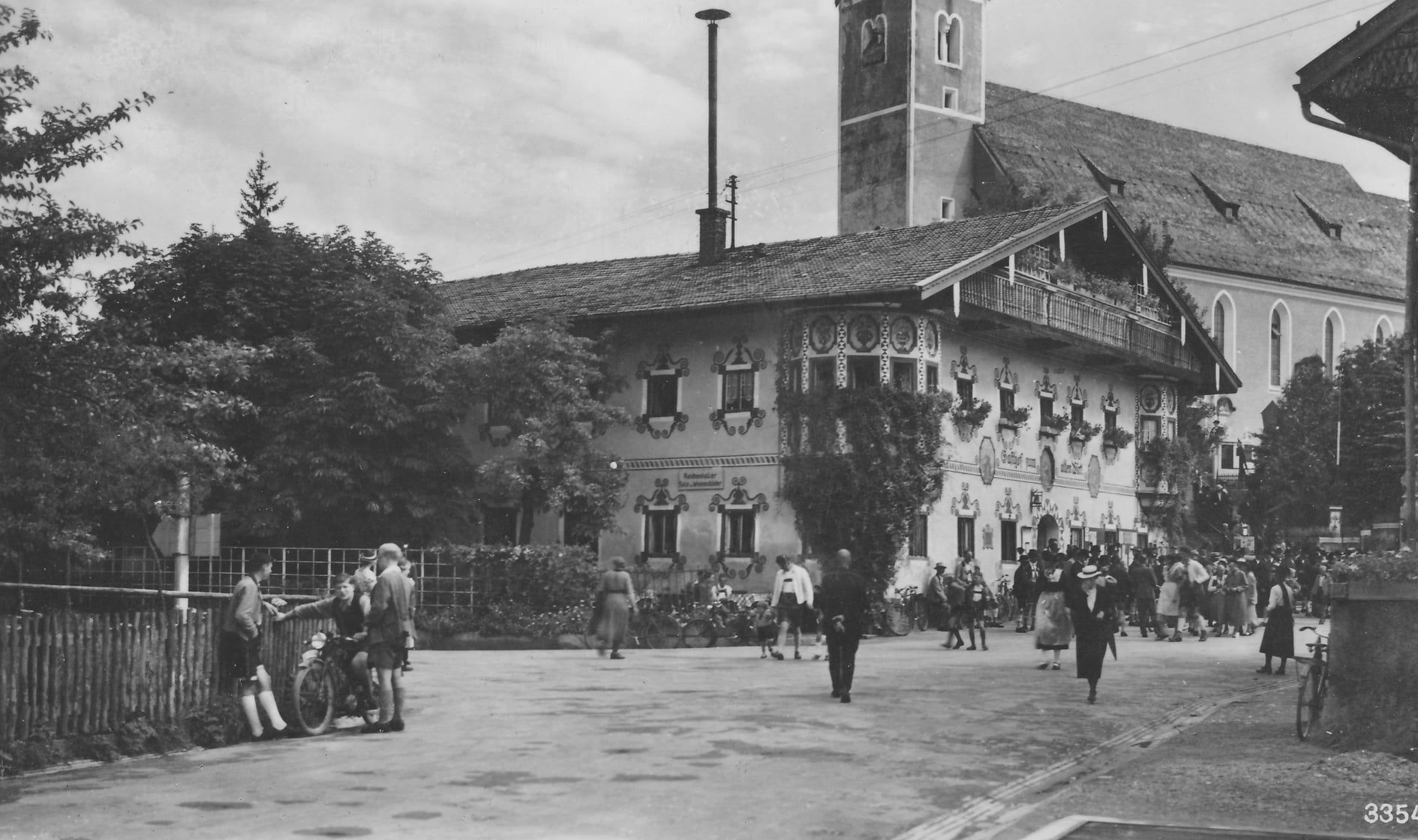 Schwarz-weiss-Aufnahme. Hintergrund Kirche, davor ein Bauernhaus. Auf der Dorfstraße gehen viele Menschen in Tracht. Links am Zaun stehen ein paar junge Burschen in Lederhosen mit einem Motorrad.