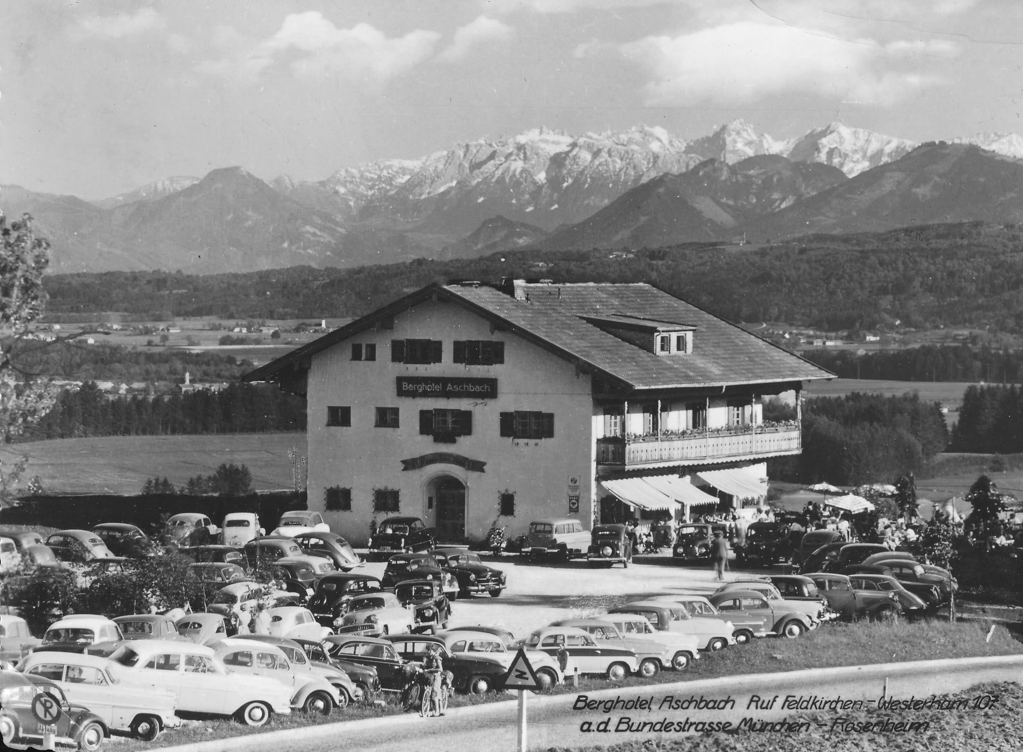 Gasthof mit Bergkulisse im Hintergrund. Davor parken viele Autos, die 1952 angesagt waren. Schwarz-Weiss-Aufnahme