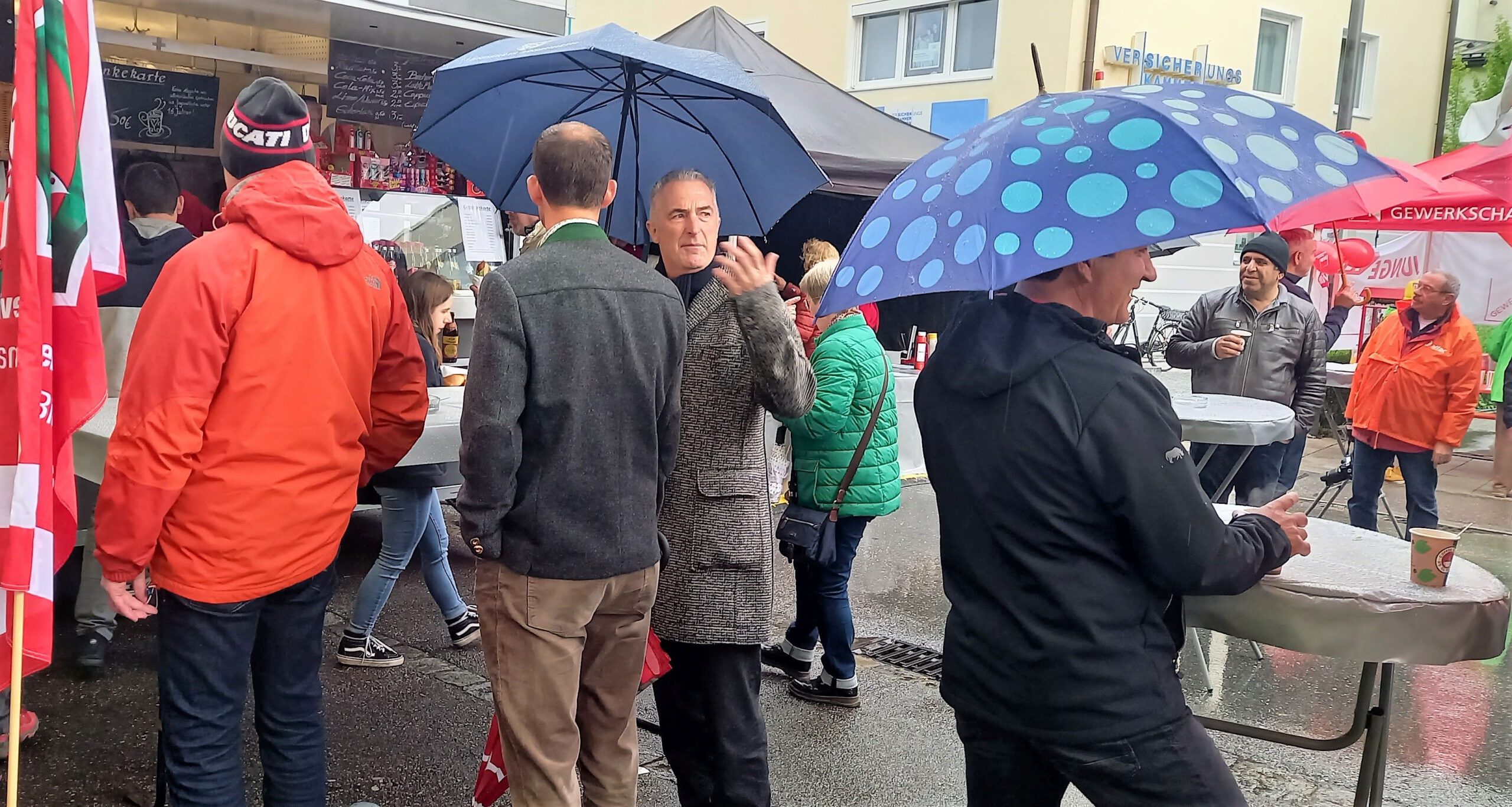 Zwei Männer sprechen mieinander. Der rechte Mann hält einen Regenschirm. Davor noch ein Mann mit Regenschirm mit großen blauen Punkten