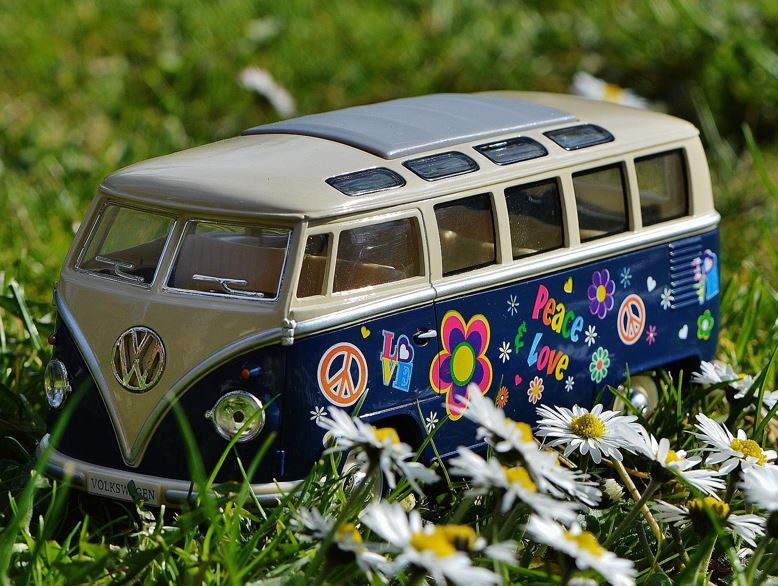 Kleiner Spielzeug VW-Campingbus, blau mit vielen bunten aufgemalten Blüten steht inmitten einer Gänseblümchen-Wiese