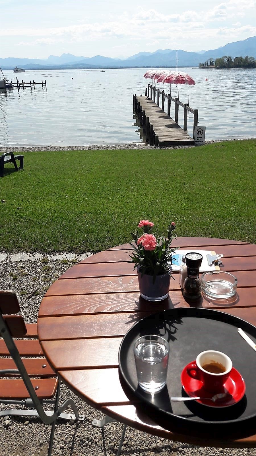 Cafè Inselblick in Gstadt am Chiemsee, Runder Tisch im Vordergrund mit Getränken darauf, dahinter Blick zum See mit Steeg und Bergkulisse