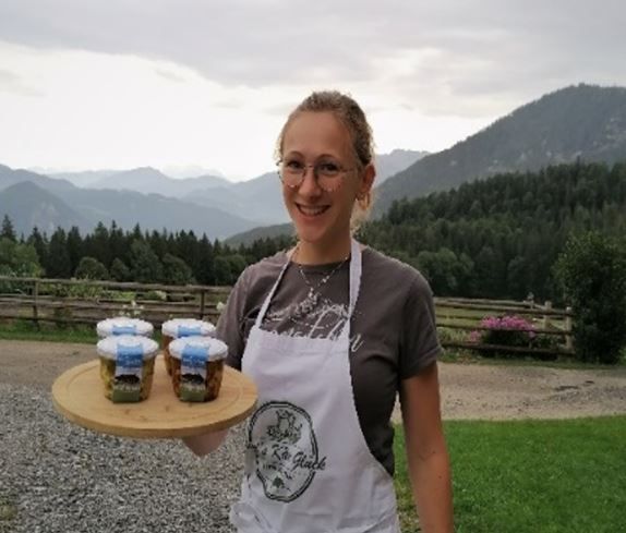 Junge Dame mit weißer Schürze hat auf Tablett vier Gläser mit selbstgemachten Käse. Im Hintergrund die Bergwelt von Bayern