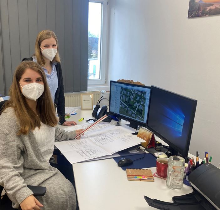 Zwei Frauen mit Mundschutzmasken, langen Haaren sitzen vor zwei Computerbildschirmen, vor sich Skizzen, Bebauungspläne.