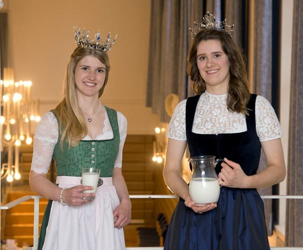 Zwei junge Damen mit Dirndl und Krone, die linke Dame mit langen blonden Haaren und die Dame rechts mit langen braunen Haaren. Links hält ein Glas mit Milch in der Hand, Rechts den Milchkrug