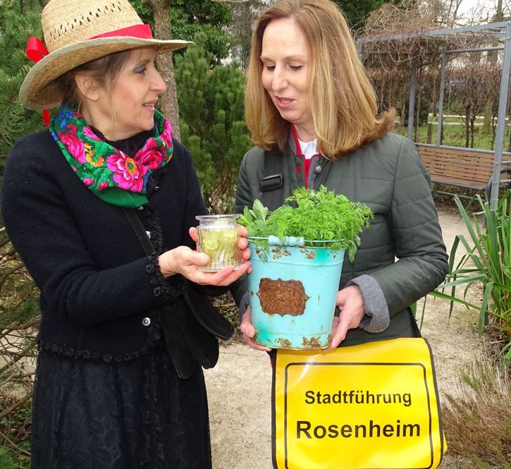 Zwei Frauen unterhalten sich im Riedergarten, rechte Dame mit Hut und Kerze in den Händen, linke Dame mit einem Kräutertopf und der gelben Stadtführung-Rosenheim-Tasche