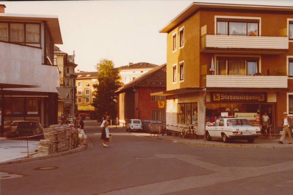 Farbaufnahme aus dem Jahr 1979. Zu sehen Blick in eine Straße. Eine Frau in Dirndl überquert sie gerade. Auf der rechten Seite kleines gelbes Gebäude mit parkendem Oldtimer davor. .