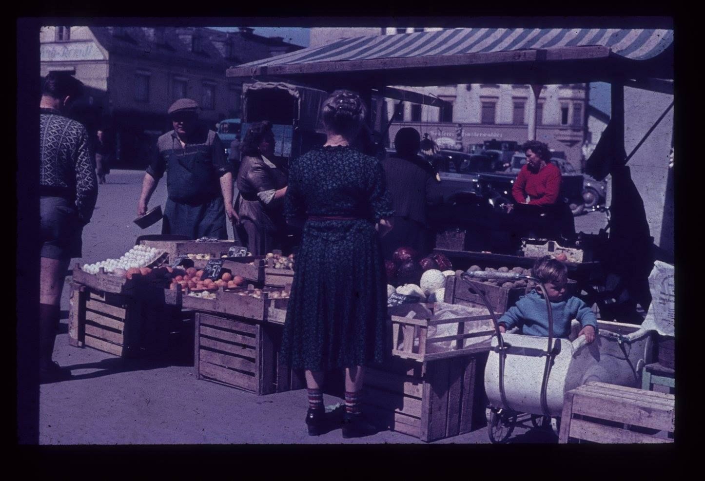 historische Farbaufnahme aus dem Jahr 1952, zeigt Szene an Marktstand. Frau in damals typischer Kleidung kauft was ein, besonderer Hingucker ist der weiße Kinderwagen der damaligen Zeit daneben mit einem sitzenden Buben mit blauem Oberteil