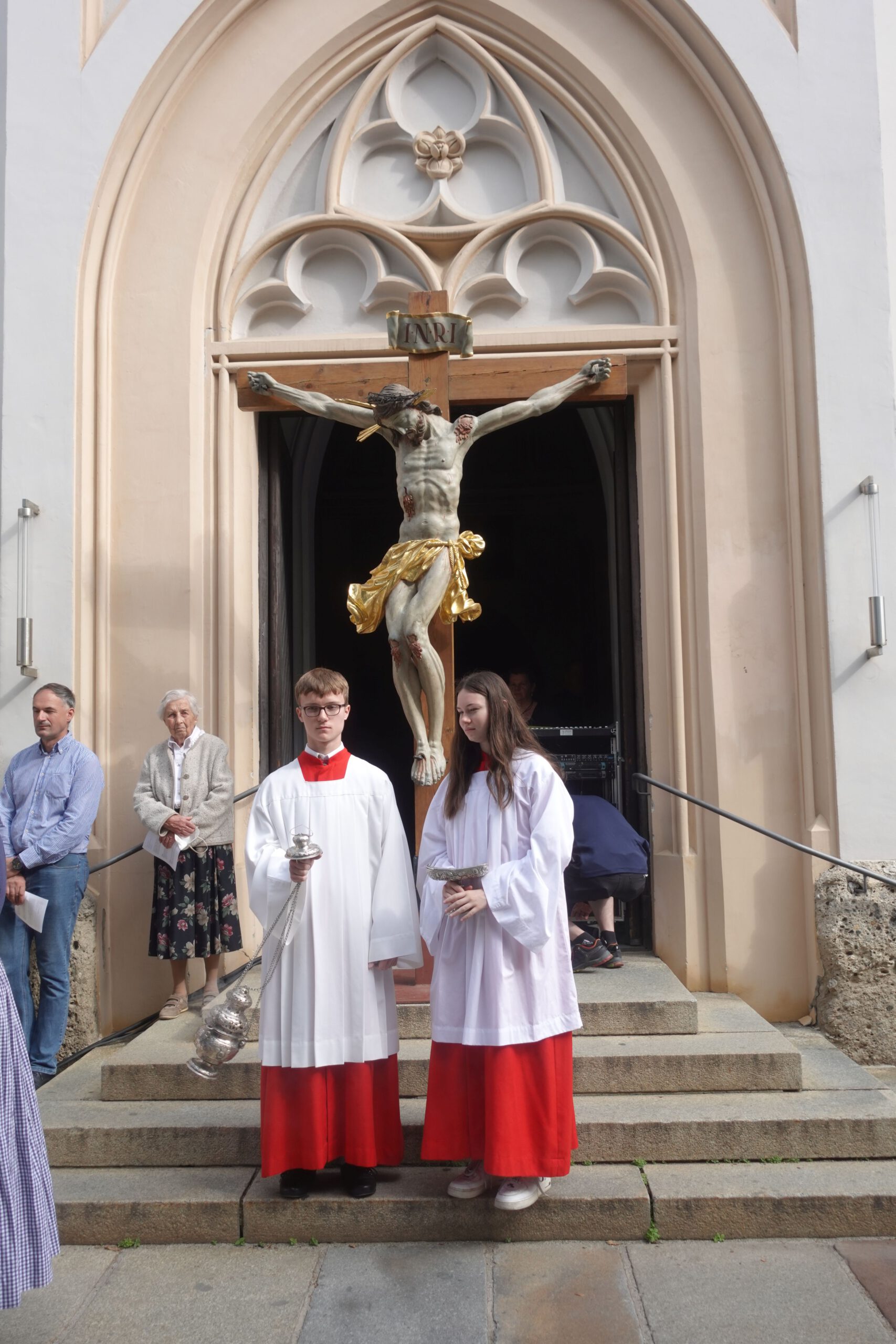 Fronleichnam Rosenheim, zwei Ministranten vor Kirche mit grossem Kreuz dahinter