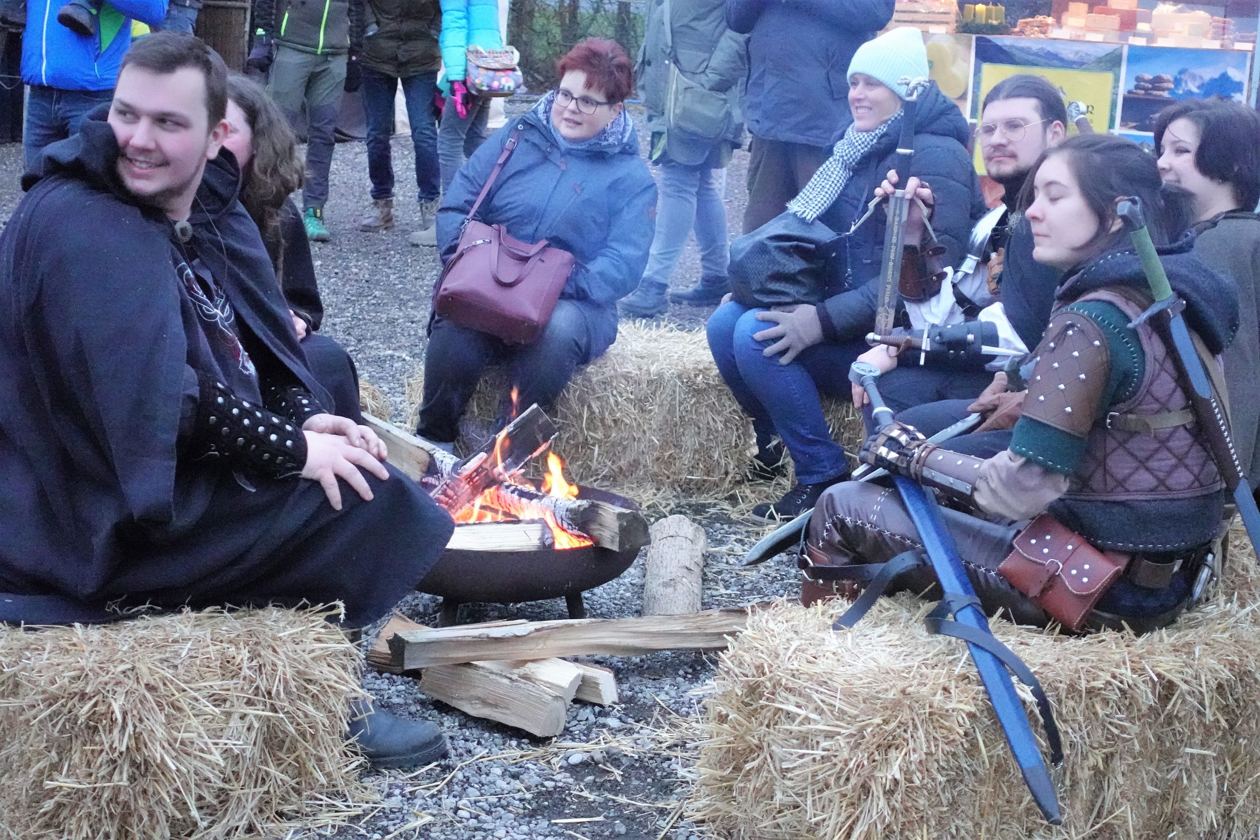 Besucher, teils mittelalterlich gekleidet, sitzen auf Heuballen um ein Lagerfeuer