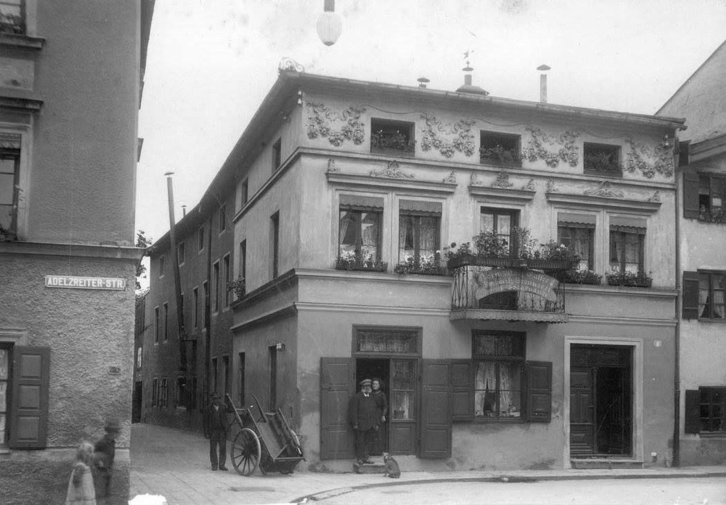Gebäude im Jahr 1911 in der Adlzeierstraße in Rosenheim mit einem Mann vor der Eingangstüre und einem Mann mit Karren in einer schmalen Gasse daneben.