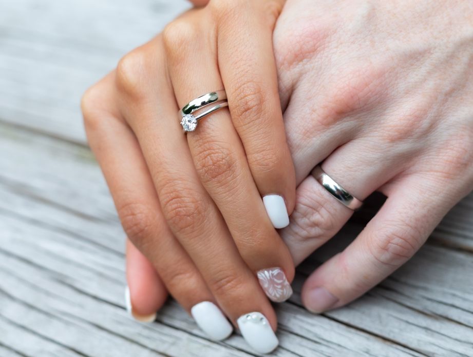 Hände eines Ehepaars mit Fokus auf die Eheringen. Sie hat weiße Fingernägel.