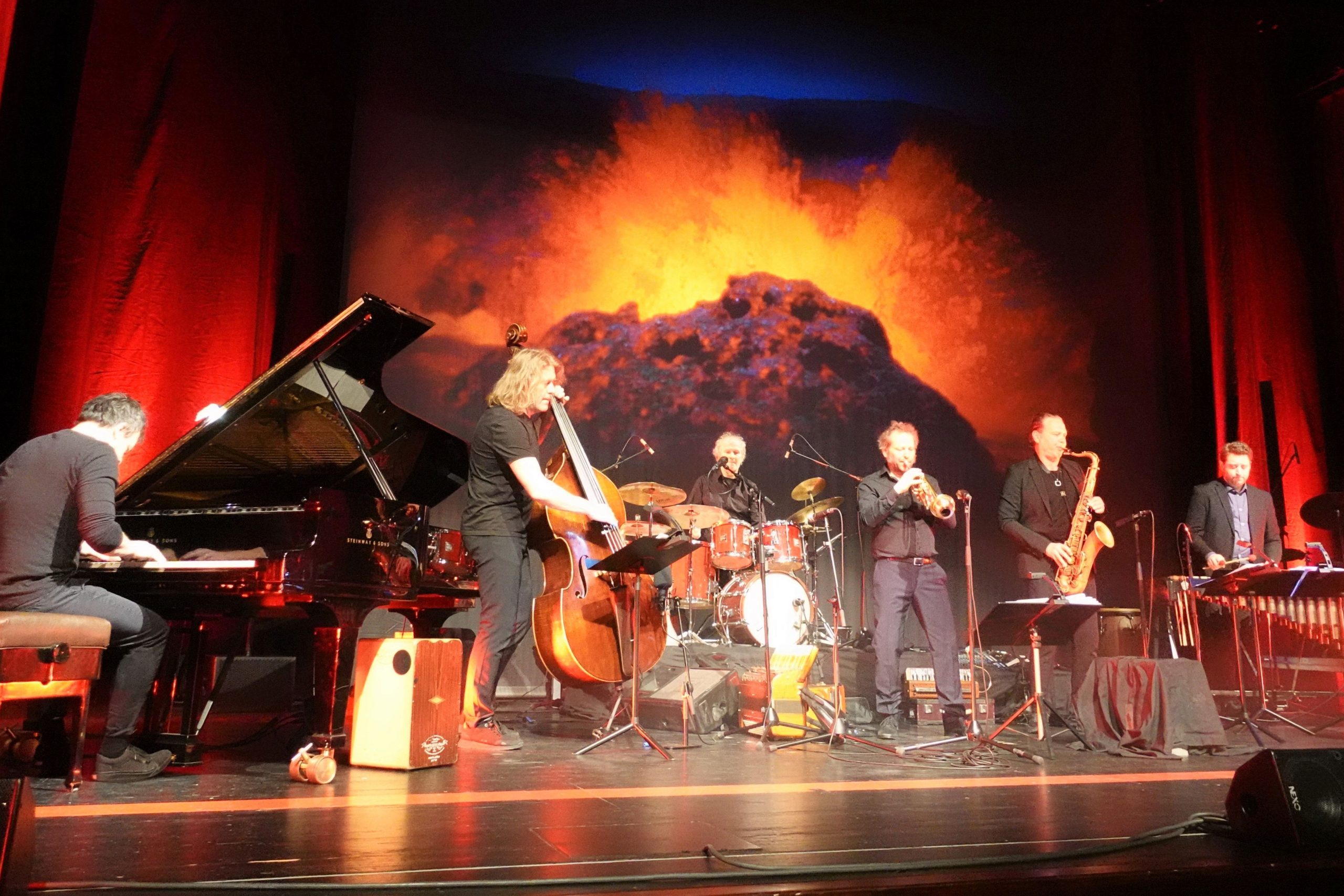 Die Band Quadro Nuevo auf der Bühne im Kultur- und Kongresszentrum Rosenheim. Dahinter auf einer Leinwand bricht ein Vulkan aus und speit Feuer und Lava in die Luft