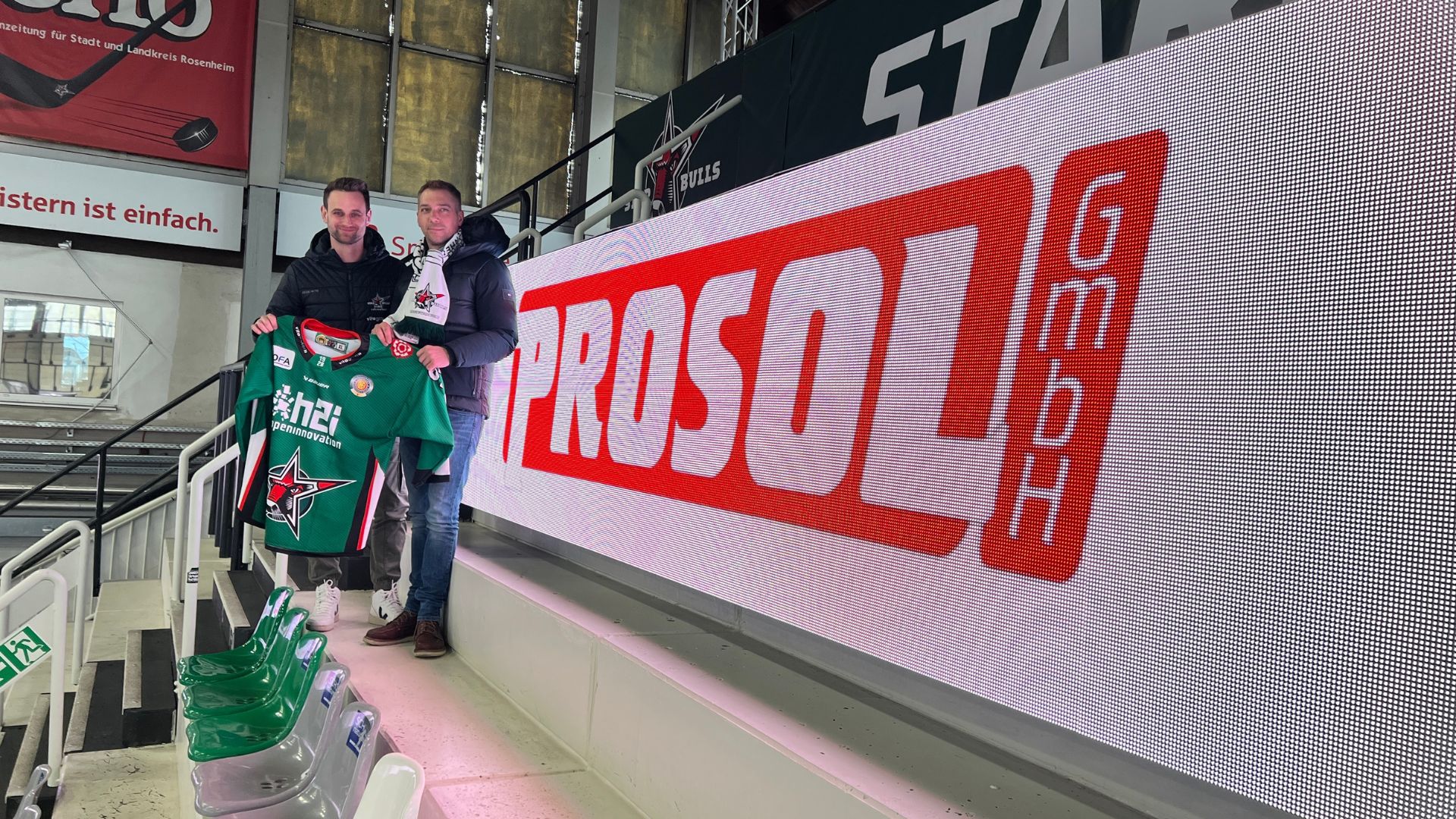 Im Rosenheimer Eisstadion mir Prosol-Banner an der Seite. Davor zwei Männer mit Starbulls-Shirt in den Händen