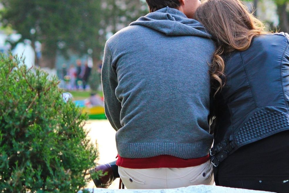 Zwei verliebte jungen Menschen sitzen auf einer Bank. Fotografiert von hinten. Auf der linken Seite ein grüner Busch.
