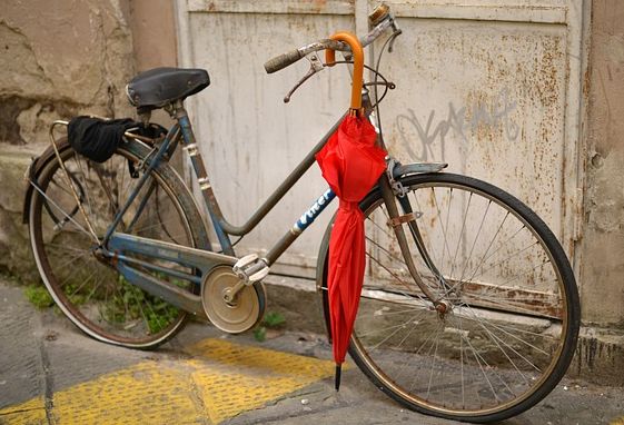 Fahrrad lehnt an der Wand. Daran angelehnt ein roter, geschlossener Regenschirm.