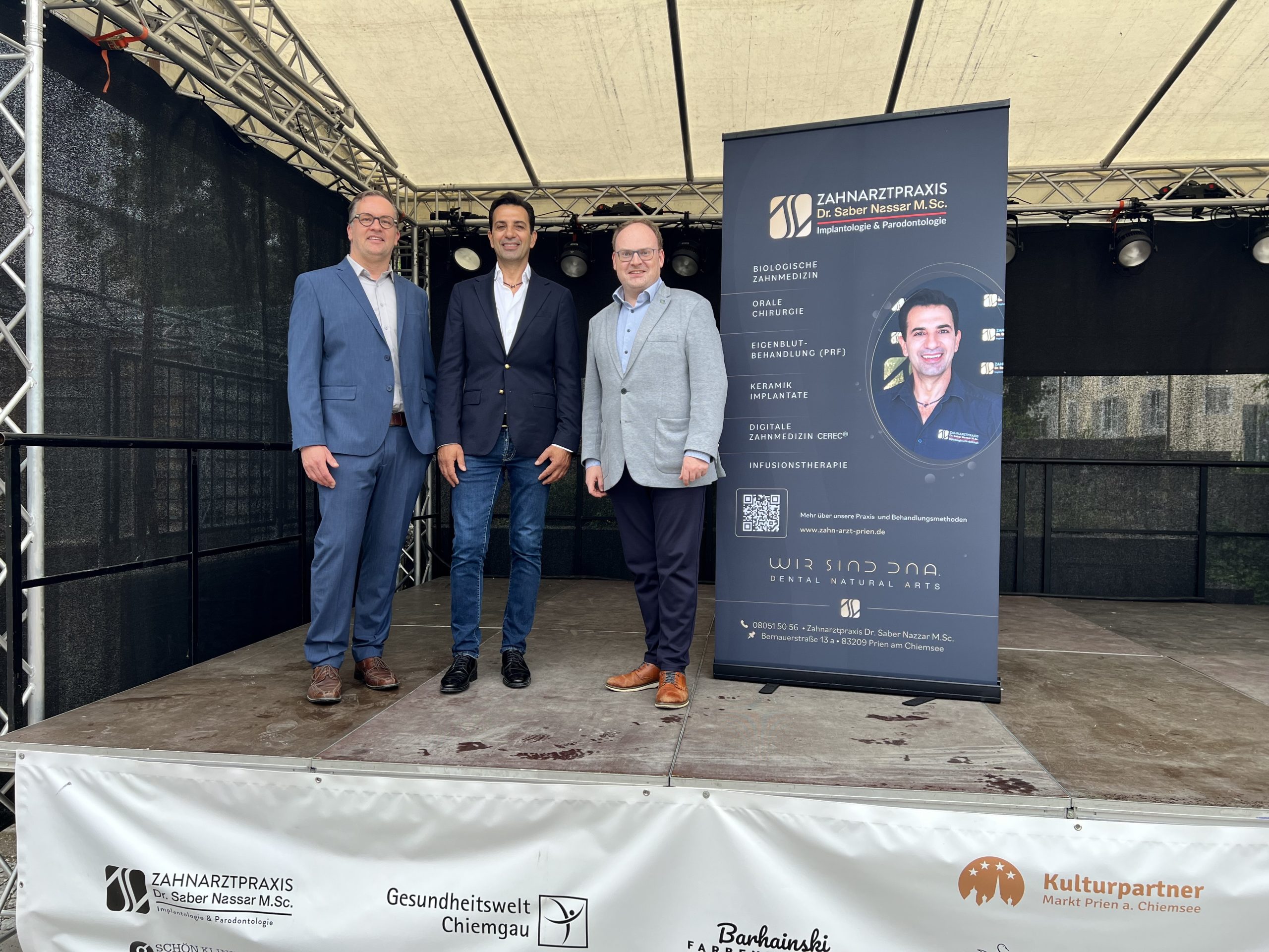 2026-07-28 Pressebild Kulturpartner c-Markt Prien a. Chiemsee Ab Oktober 2026 setzt die Zahnarztpraxis von Dr. Saber Nassar M.Sc. (im Bild mittig) als neuer Platin-Partner ein sichtbares Zeichen für ihr Engagement im kulturellen Leben Priens. Gemeinsam mit dem Ersten Bürgermeister der Marktgemeinde, Andreas Friedrich (rechts), stellt der Fachbereichsleiter für Kultur, Tourismus und Wirtschaftsförderung Tobi Huber (links) die neue Partnerschaft vor. Foto: Copyright Markt Prien